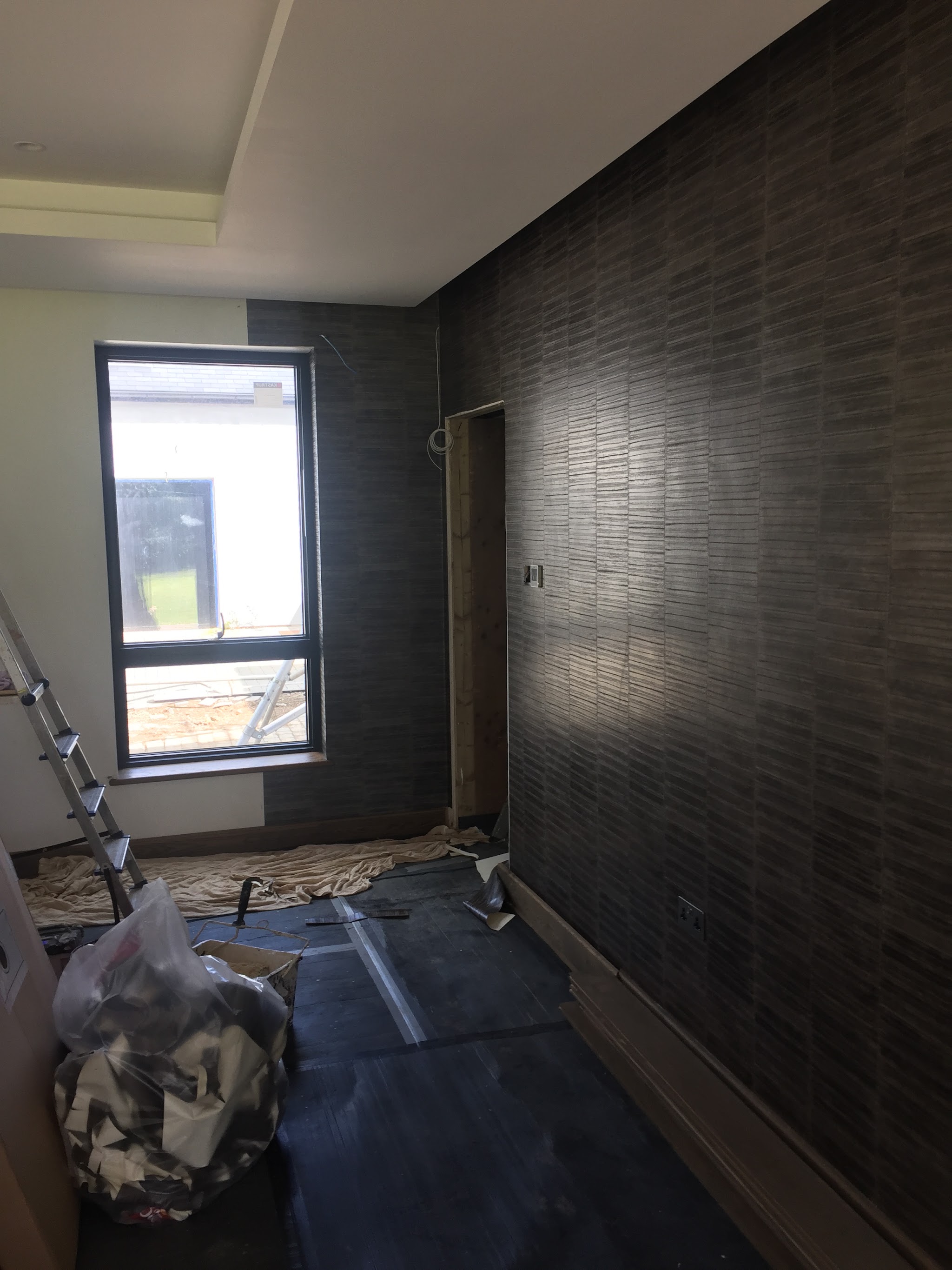 Long wall being papered in a dark textured feature wallpaper, with dust sheets and a step ladder